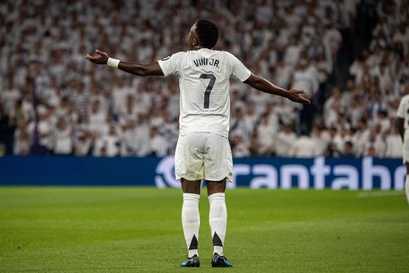 Soccer player in a white jersey with Vinicius 7 stands with arms outstretched on a green field, facing a blurred crowd.