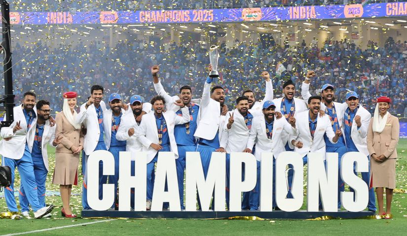 A cricket team in blue and white celebrates a victory, holding a trophy with confetti falling. They stand behind large CHAMPIONS letters.