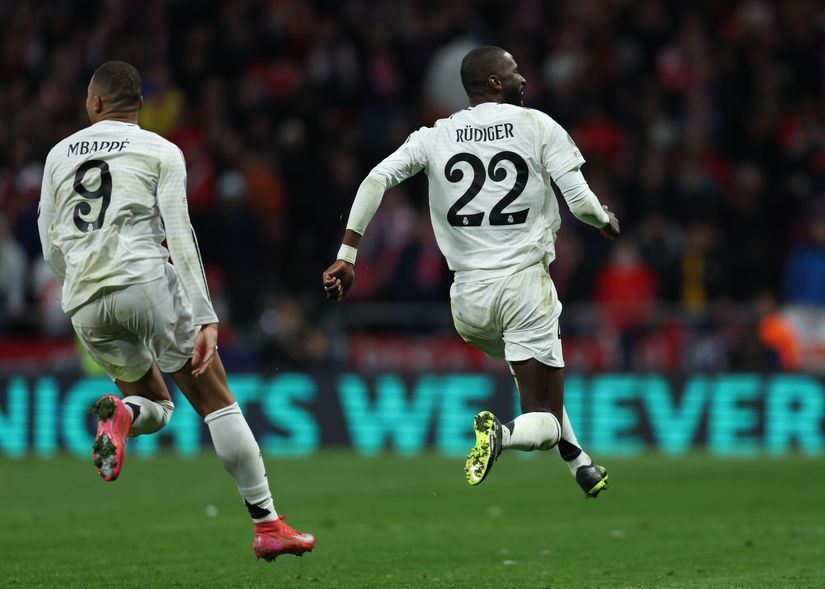Two soccer players in white jerseys are running on a field. The players jerseys display numbers 9 and 22. The background shows a blurred crowd and part of a large, partially visible sign.
