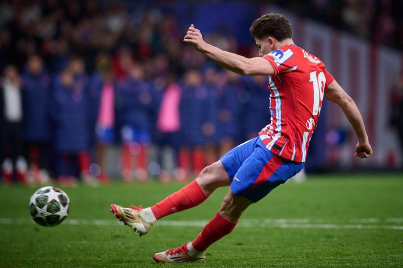 Soccer player in red and white jersey kicking a ball on a field during a match, with spectators in the background.