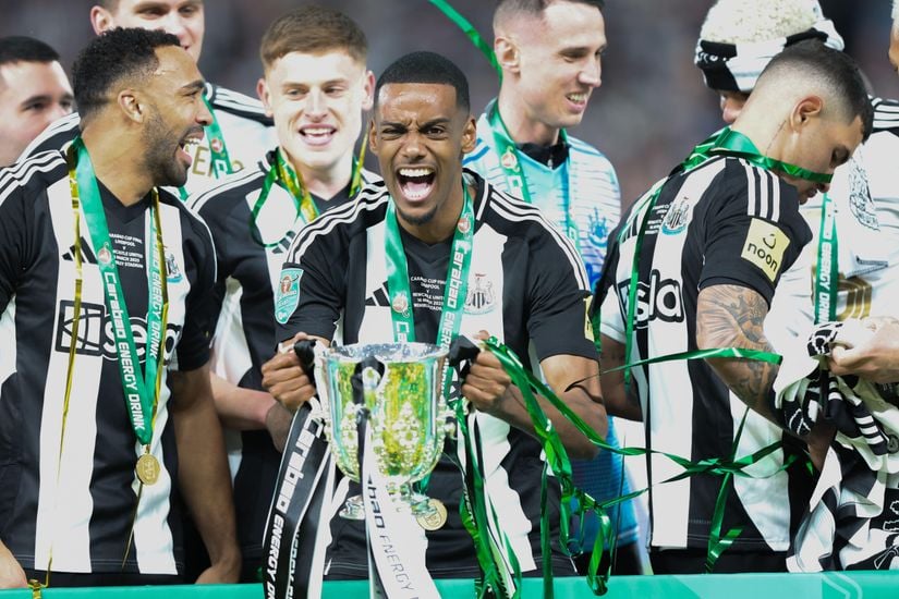 Soccer players celebrating with a trophy. Player in front holding the trophy and shouting. Green and black ribbons dangle from the cup. Others behind are smiling and wearing medals.