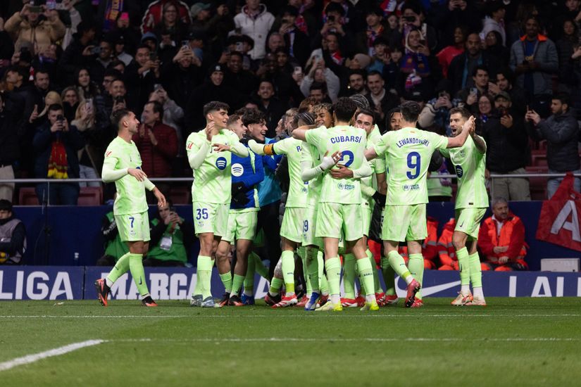 A soccer team wearing bright green uniforms celebrates on the field. Players are huddled, some with arms raised. A crowd watches in the background.
