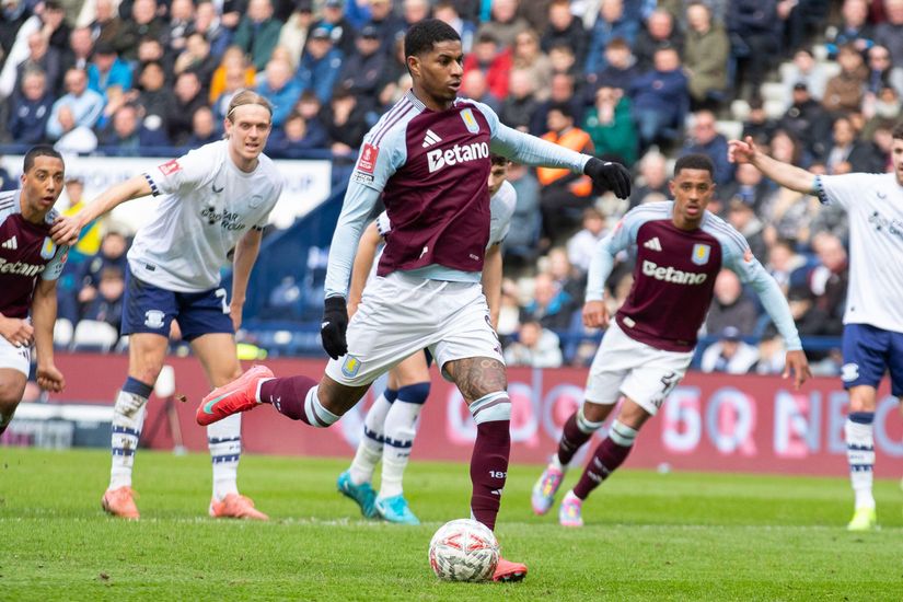 A soccer player in a maroon and light blue uniform prepares to kick the ball toward the goal during a match. Opposing team players in white and navy uniforms look on.