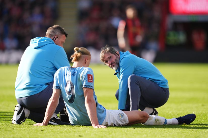 A football player in light blue sits on the pitch while two staff members in blue check on him.