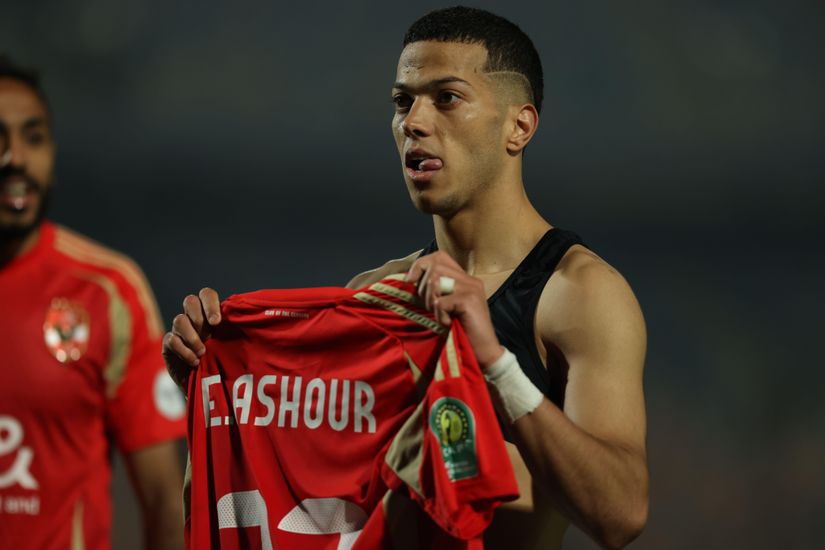 A soccer player holds up a red jersey with F. Ashour written on it, wearing a black sleeveless shirt on the field. Another player is partially visible in the background.