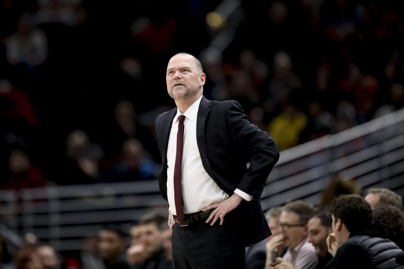 A basketball coach in a suit stands on the sidelines with a focused expression, hands on hips.