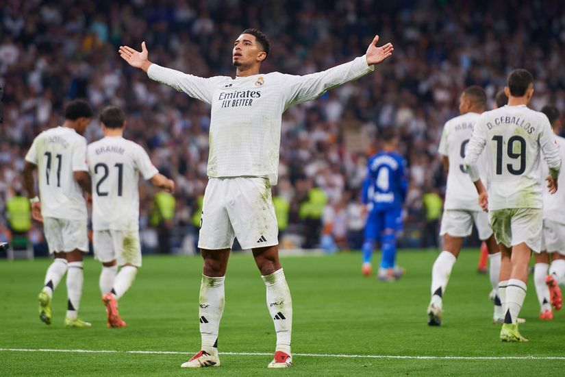 Soccer player in a white jersey stands with arms outstretched on a field, surrounded by teammates and opponents.