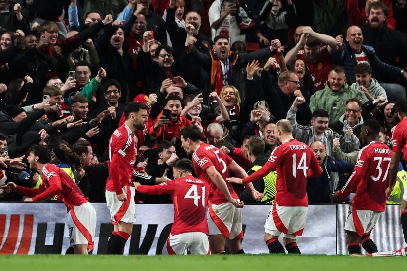 Soccer players in red jerseys celebrate a goal near the stands as cheering fans react enthusiastically in the background.