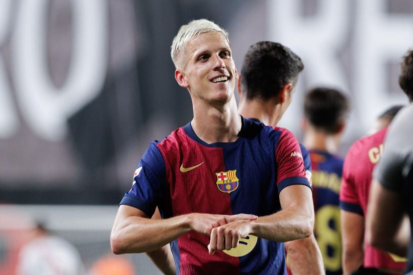 Soccer player in a Barcelona jersey stands on the field, smiling, with other teammates visible in the background.
