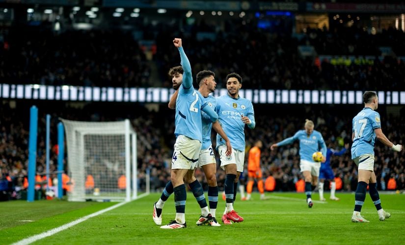 Manchester City players celebrate a goal on the pitch during a football match, with one player raising his arm and fans visible in the stadium background.
