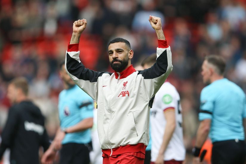 A soccer player in a Liverpool jacket raises both arms in celebration on the field, with referees and other players in the background.