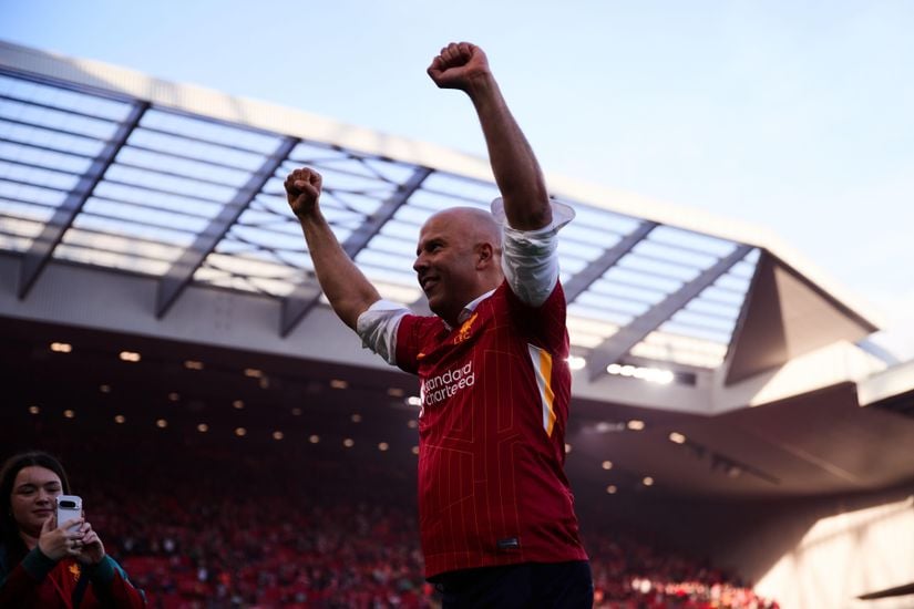 A person in a red soccer jersey stands with both arms raised in celebration on a stadium field, with a crowd and open roof structure in the background.