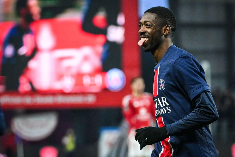 A soccer player in a PSG jersey smiles and sticks out his tongue on the field, with a blurred stadium screen and crowd in the background.