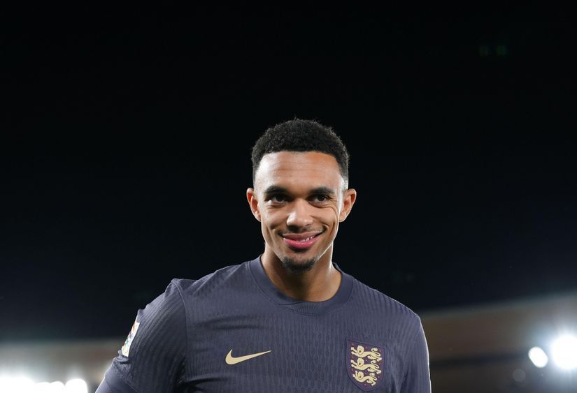 A man wearing a dark England football jersey stands smiling, with a dark background and stadium lights partially visible.