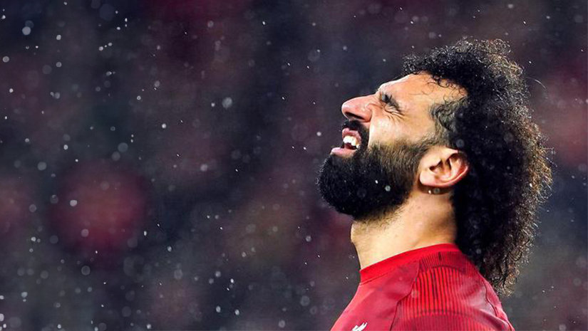A soccer player with curly hair and a beard, wearing a red jersey emblazoned with BW Arabia, looks upwards with a determined expression. Surrounded by raindrops against a dark background and blurred lights, he embodies resilience and focus.