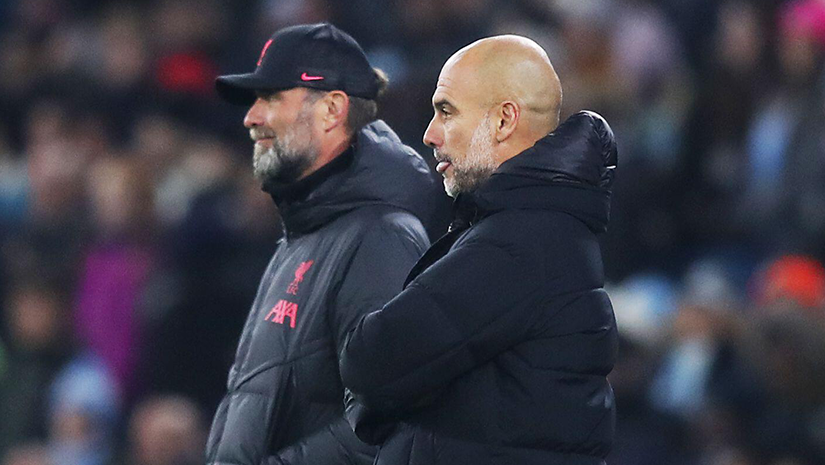 Two soccer coaches stand side by side on the sidelines, both wearing dark jackets. One wears a cap, and the other is bald. They appear focused, watching the match intently with spectators blurred in the background, embodying the competitive spirit of Betway Arabia.