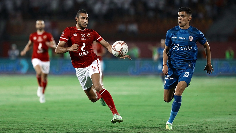 لاعبان لكرة القدم، أحدهما يرتدي قميصًا أحمر والآخر أزرق، يطاردان الكرة على ملعب أخضر. يبدو أن اللاعب الذي يرتدي قميصًا أحمر يركل الكرة بينما يركض اللاعب الذي يرتدي قميصًا أزرق بجانبه. يهتف مشجعو بيتواي أرابيا من جمهور غير واضح في الخلفية.