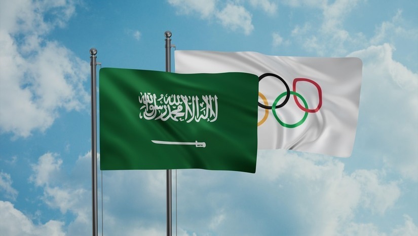 Two flags side by side against a cloudy sky. The left flag, symbolizing Saudi Arabia, is green with white Arabic script and a sword. The right flag is white with the five iconic Olympic rings. This scene captures the spirit of unity much like the global community found in bw Arabia events.