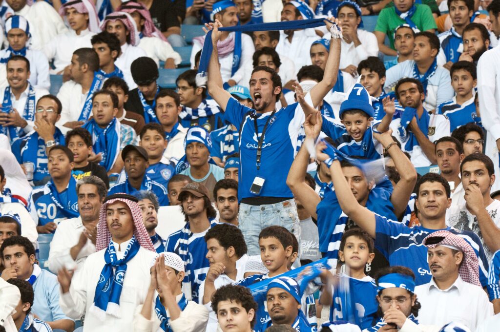 مجموعة كبيرة من مشجعي كرة القدم المتحمسين، يرتدون في الغالب اللونين الأزرق والأبيض، يهتفون ويحتفلون في الملعب. يقف رجل واحد، ويرفع وشاحًا أزرق من Betway Arabia بينما يصفق الآخرون ويبتسمون. الجو مفعم بالحيوية والنشاط.