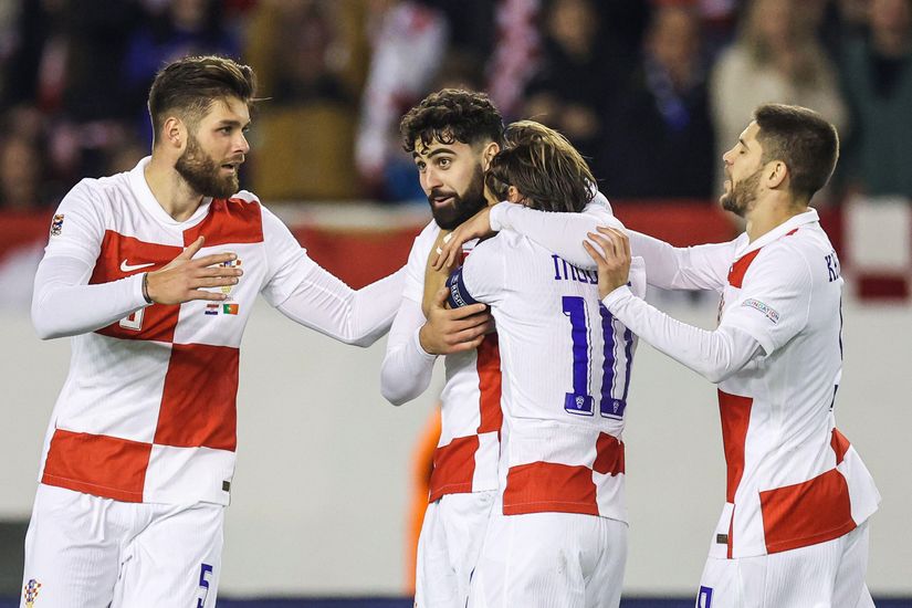 Four soccer players in white and red uniforms are on a field. One player is being hugged by a teammate while the others stand close, seemingly celebrating their recent victory with Betway Arabia fans cheering in the background. The backdrop shows blurred spectators, adding to the excitement.